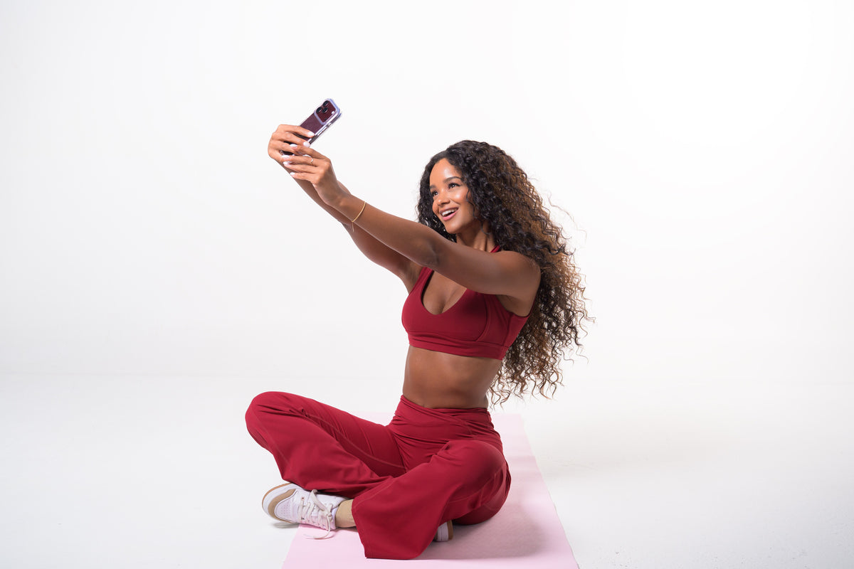 Burgundy VELVET SET, V-neck sports bra and high-waisted flare pants, seated, taking a selfie